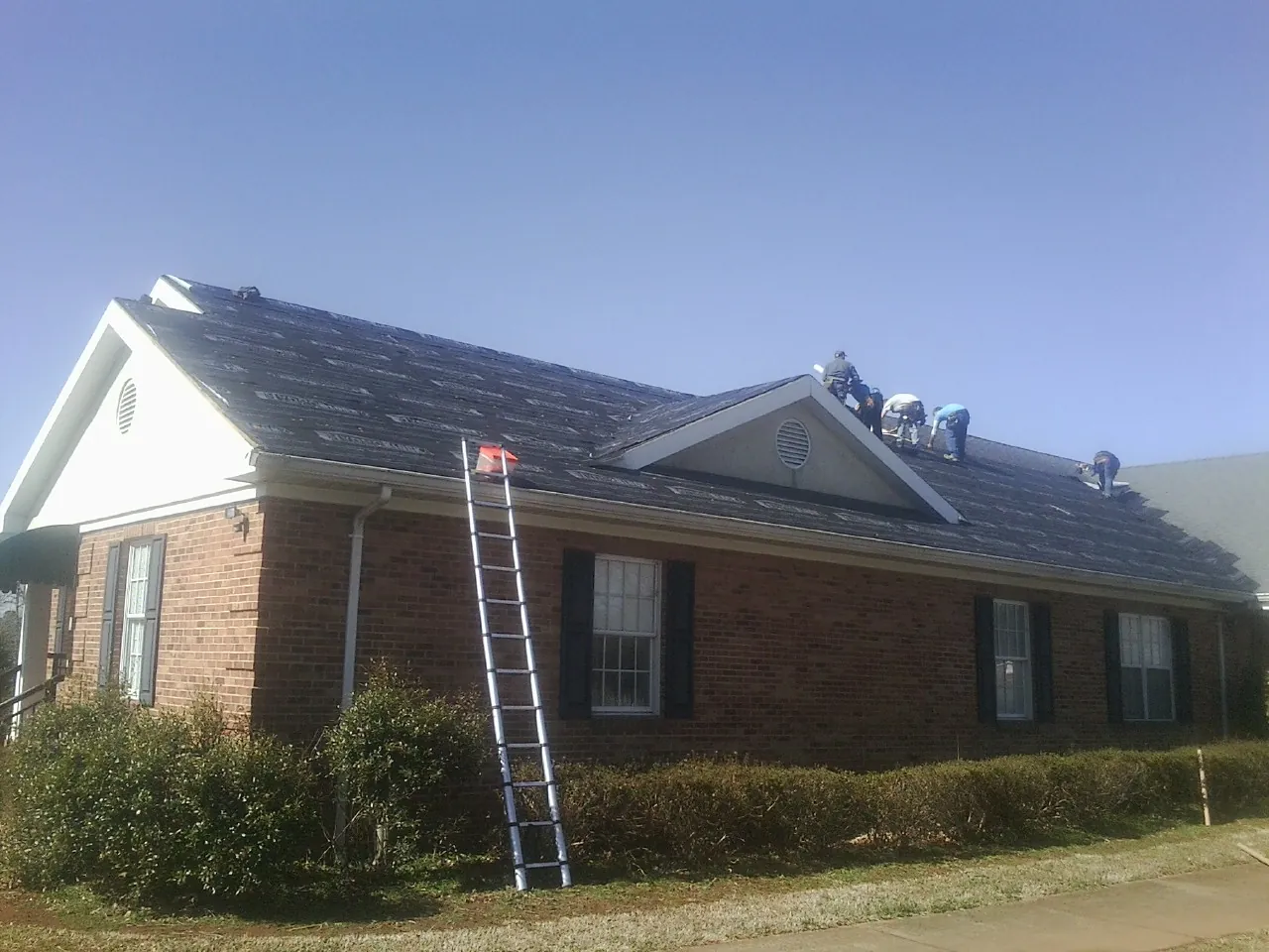 Crew tearing shingles off a brick ranch roof with a ladder set — Metal Roof Repair work by our crew in Pataskala