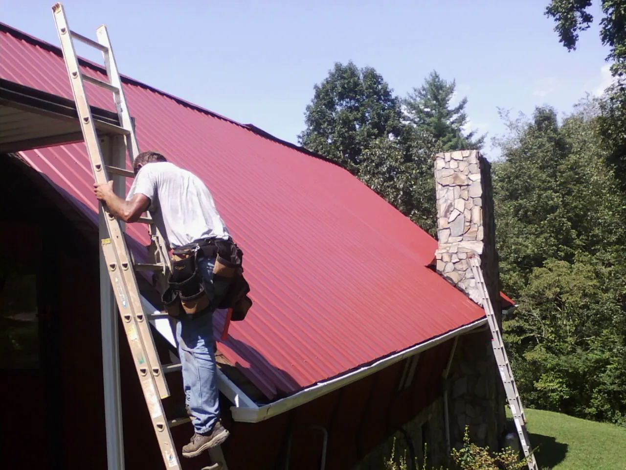 Roofer on a ladder beside a red metal roof and stone chimney — Soffit & Fascia Repair by Roofing Brothers in Pataskala